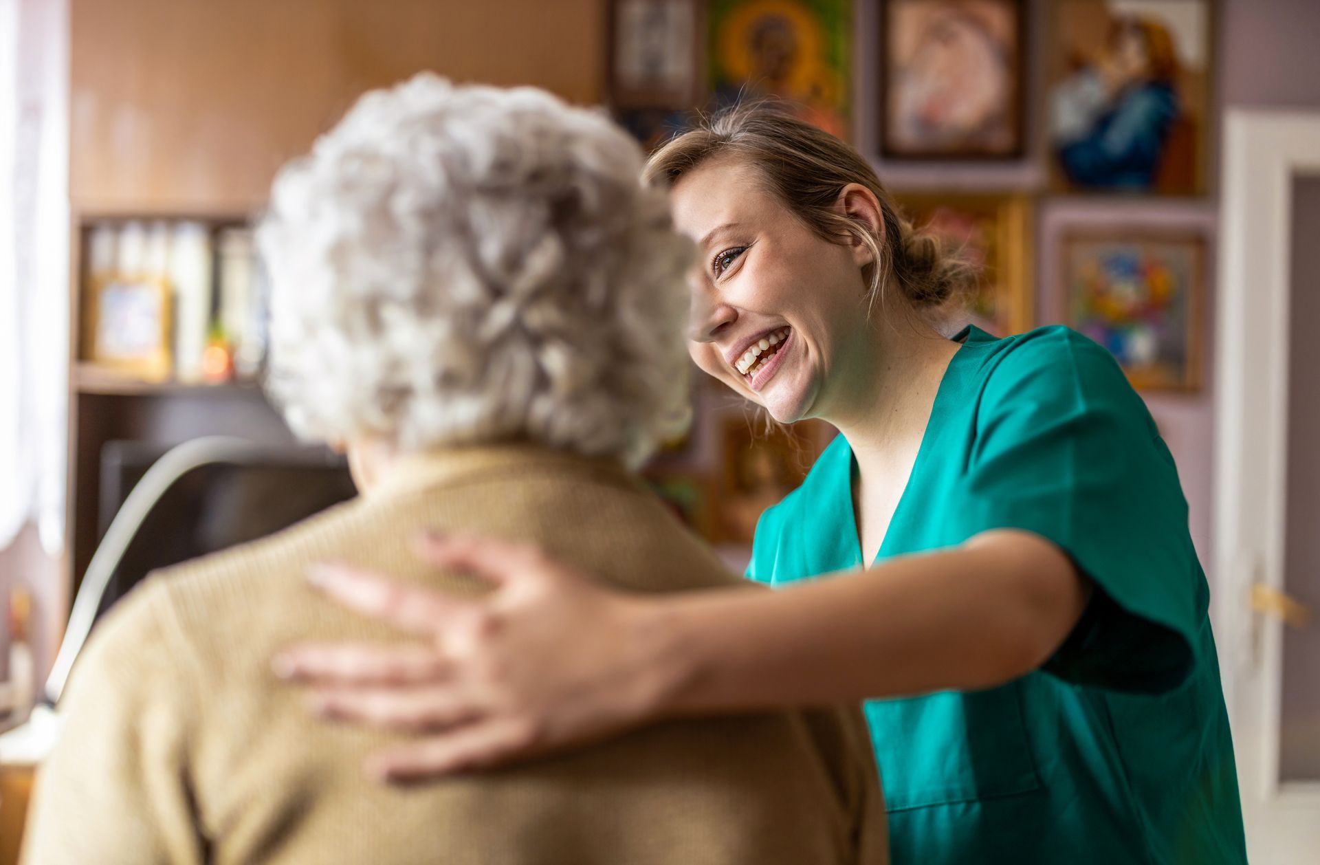 A smiling caregiver comforts an older person, placing a hand on their shoulder indoors.
