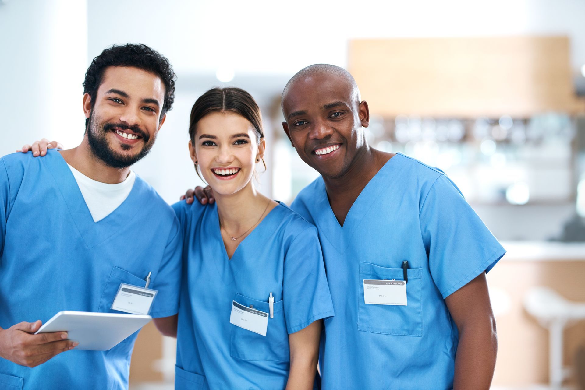 Three healthcare professionals in blue scrubs smiling, in a bright hallway.