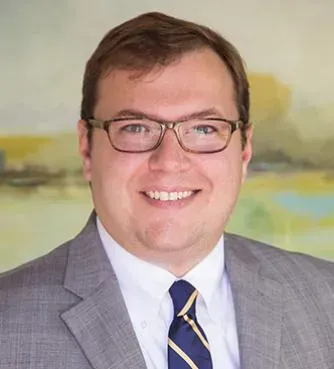 Smiling man in glasses wearing a gray suit and striped tie against a soft abstract background