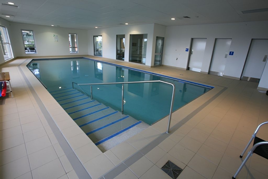 Indoor pool with steps and handrails, light tile floor, white walls, blue water. — Banora Pools in Tweed Heads South, NSW