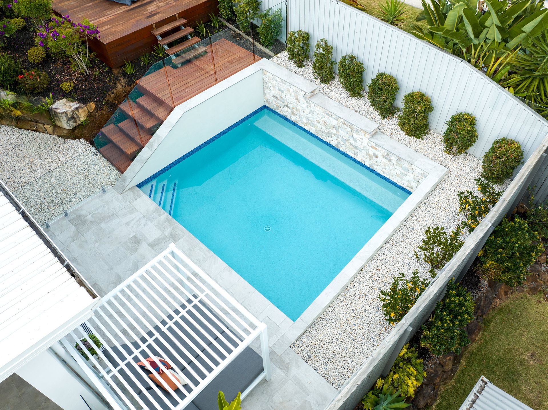 Aerial view of a small, blue swimming pool surrounded by landscaping and a wooden deck.
