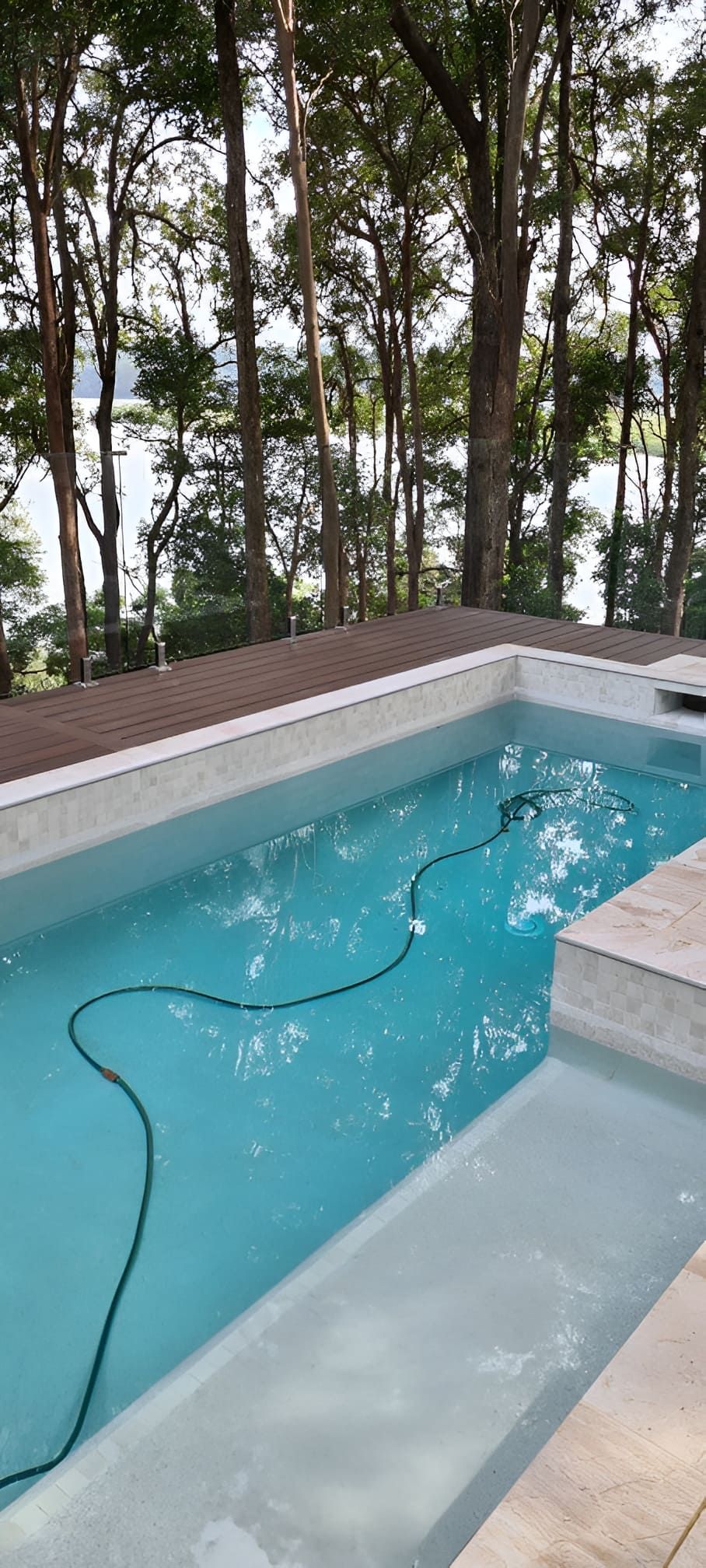 A rectangular pool with clear blue water, a black hose, surrounded by trees and a lake view. — Banora Pools in Mullumbimby, NSW