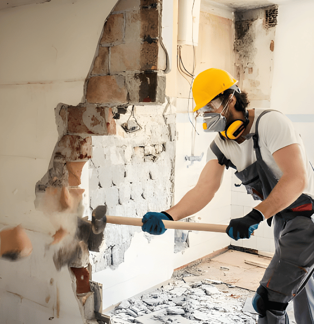 Construction worker in safety gear demolishing a wall with a sledgehammer. Debris covers the floor.