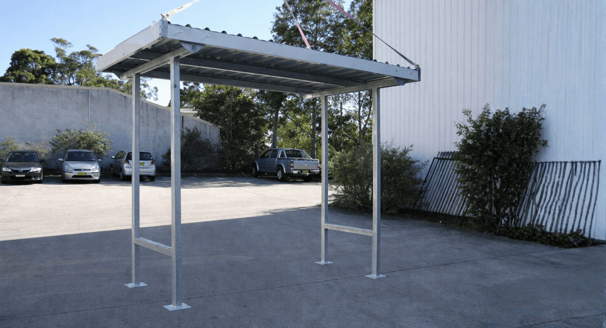 A freestanding, gray metal shelter structure with a flat roof stands in an outdoor parking lot with cars in the background.
