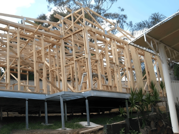 A house under construction with a wooden frame elevated on steel posts, set against a background of trees and blue sky.