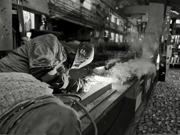A person in protective gear welding metal in a workshop, surrounded by sparks and smoke in a black-and-white setting.