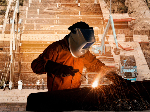 A welder in protective gear and a helmet works on a metal project, creating bright orange sparks at a construction site.