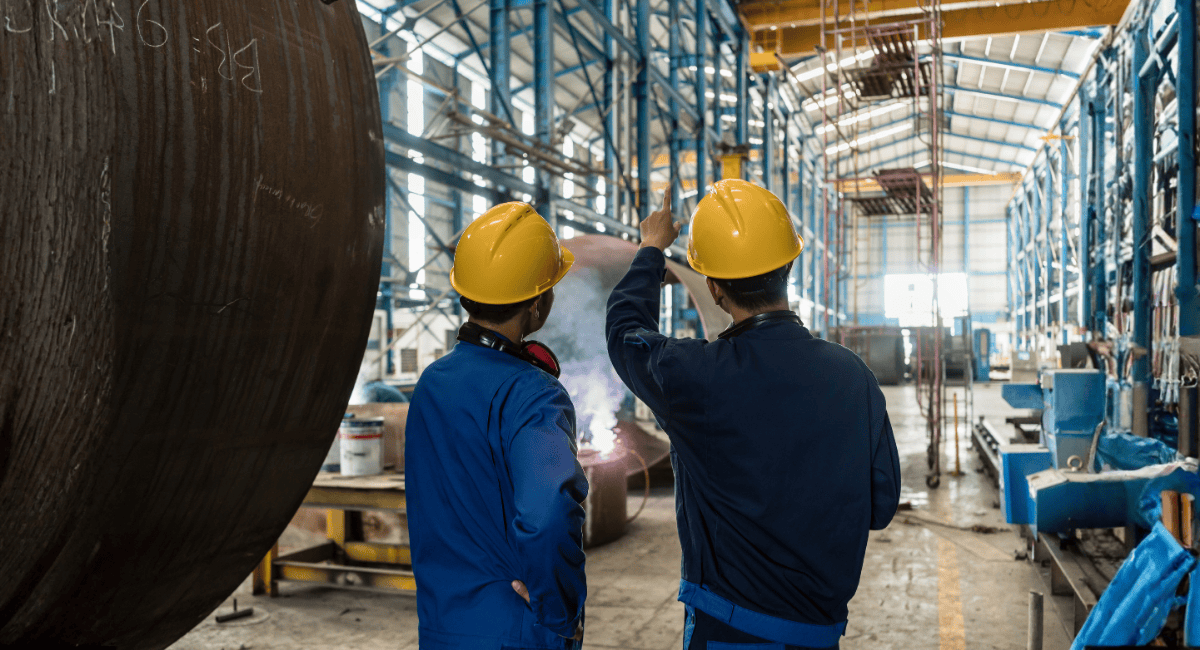 Two workers in hard hats and blue uniforms stand in a factory, one pointing toward welding work being performed.