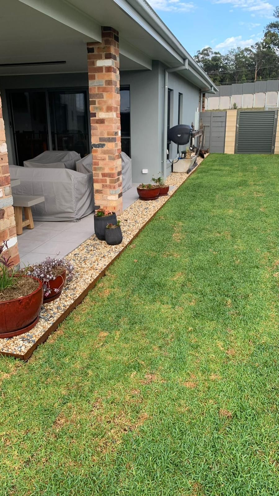 A covered patio with a brick pillar, gravel border, and potted plants next to a grassy backyard under a clear blue sky.