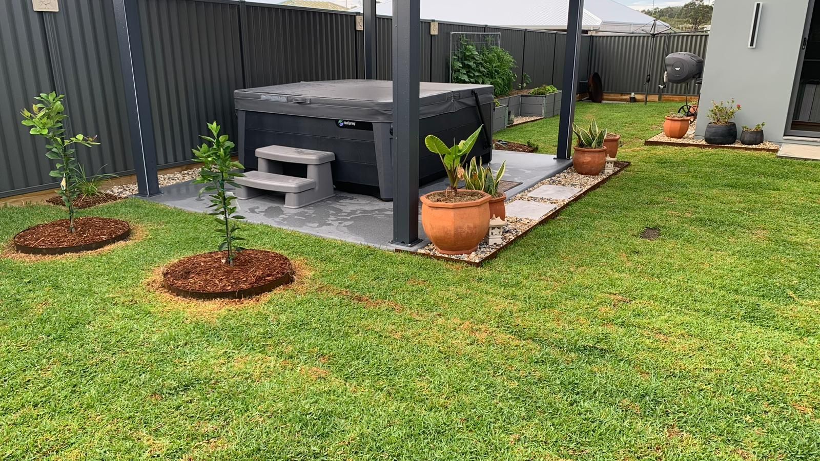 A dark spa with grey steps on a concrete patio in a backyard with a grass lawn, potted plants, and young trees.