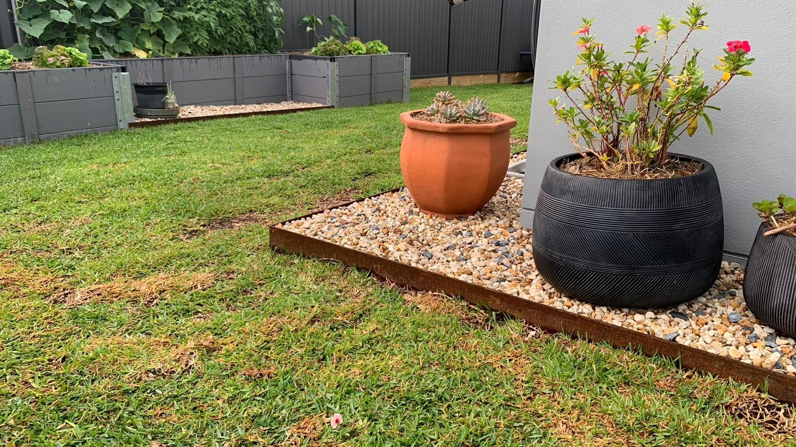 A backyard garden with raised grey planters, gravel borders with steel edging, and potted plants on a patio.