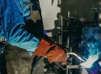 A welder in blue coveralls and orange protective gloves uses a torch to weld metal, creating a bright arc of light.