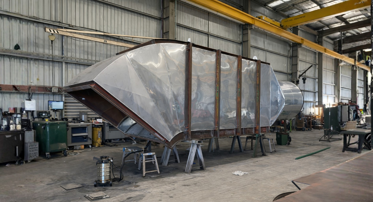 A large, industrial metal duct piece rests on supports inside a fabrication warehouse.