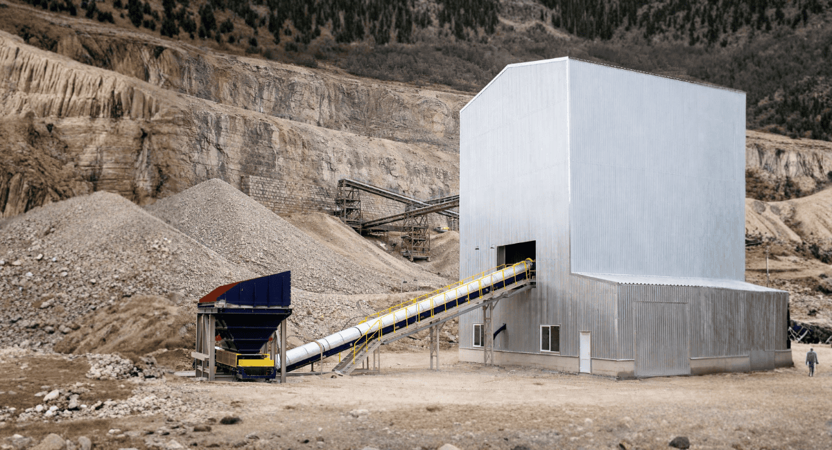 A conveyor belt transports material into a large metal structure at a quarry set against a rocky hillside.