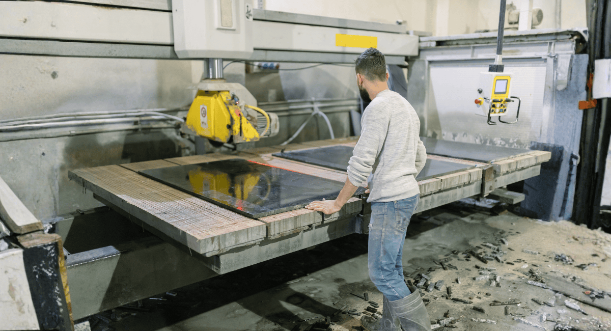 A person uses a bridge saw machine to cut a large slab of dark stone in a workshop.
