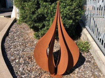 A weathered, rust-colored metal sculpture with a flame-like shape stands in a garden bed with rocks and a green shrub.