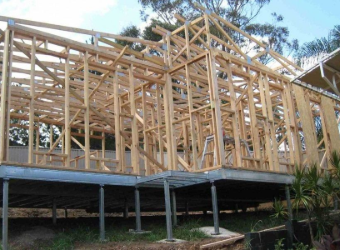 A wooden house frame under construction, supported by metal stilts on a sloping, grassy site with trees in the background.