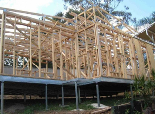 A wooden house frame under construction, supported by metal posts on a sloped site with trees in the background.