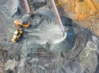 Aerial view of a yellow loader at a mining site loading dark material from a conveyor belt pile.