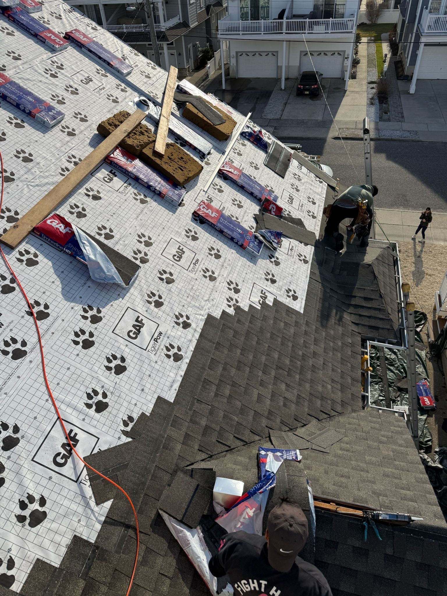 Roofing work in progress on a patterned white roof, with workers and tools near a stairway below.