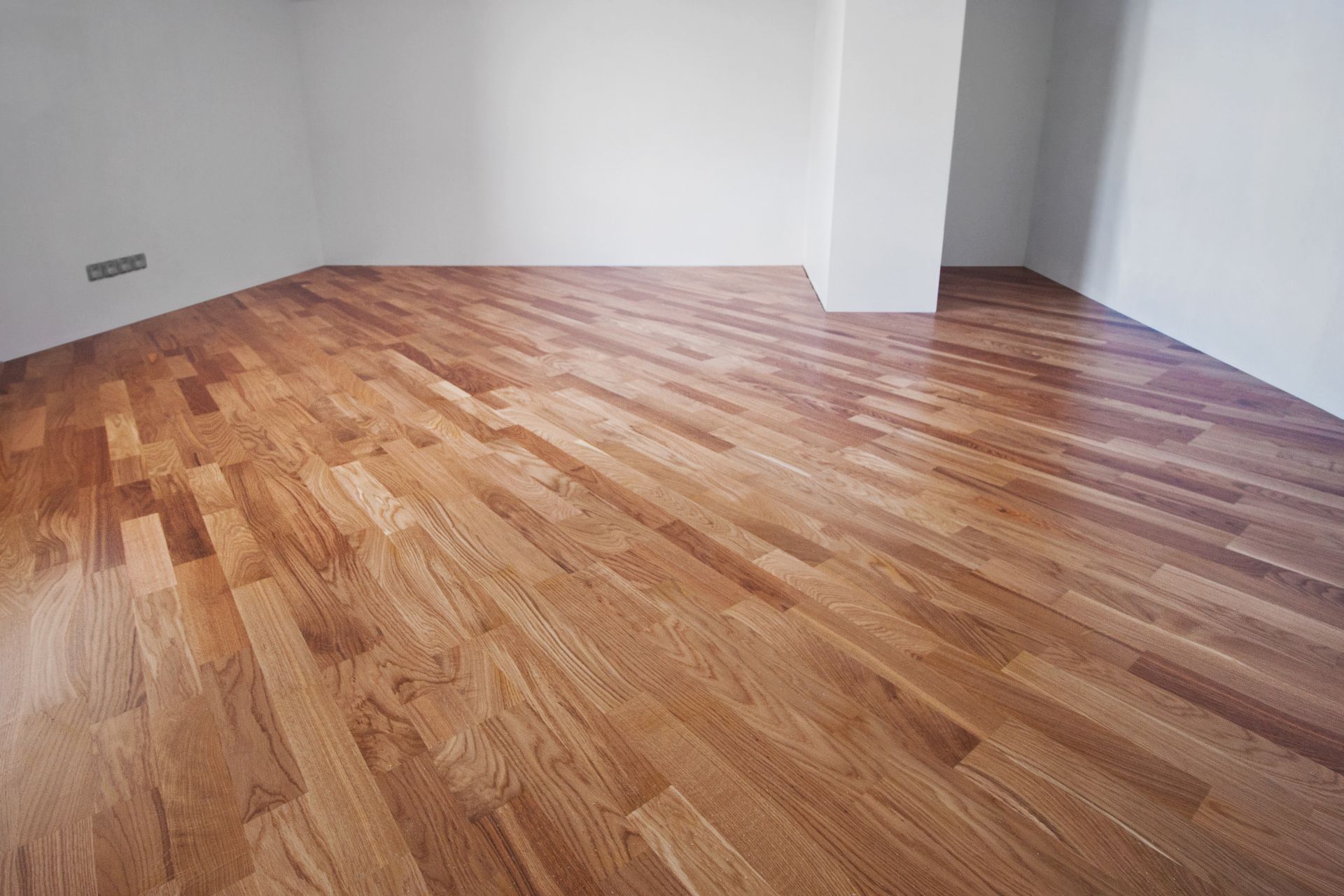 Empty room with glossy wooden floor and white walls, viewed from a corner.