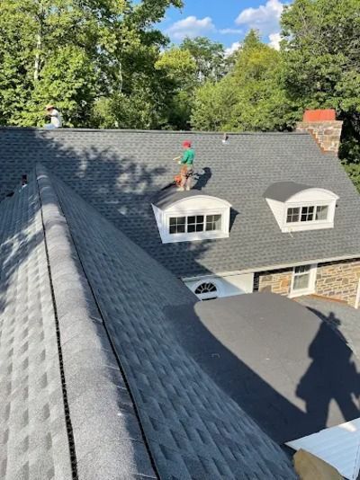 Roofer on dark shingled roof beside dormer windows, with trees and blue sky in the background