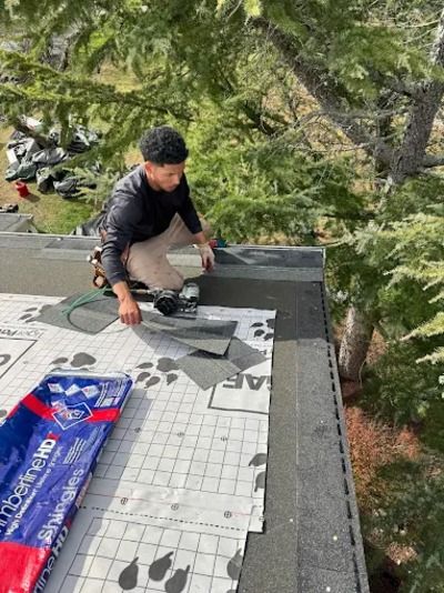 Worker installing roofing underlayment on a steep roof, with trees in the background.