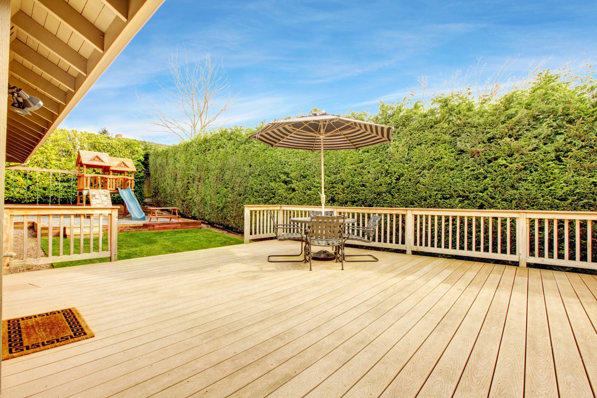 Sunny backyard deck with white railing, patio table and umbrella, and a hedge-lined garden backdrop
