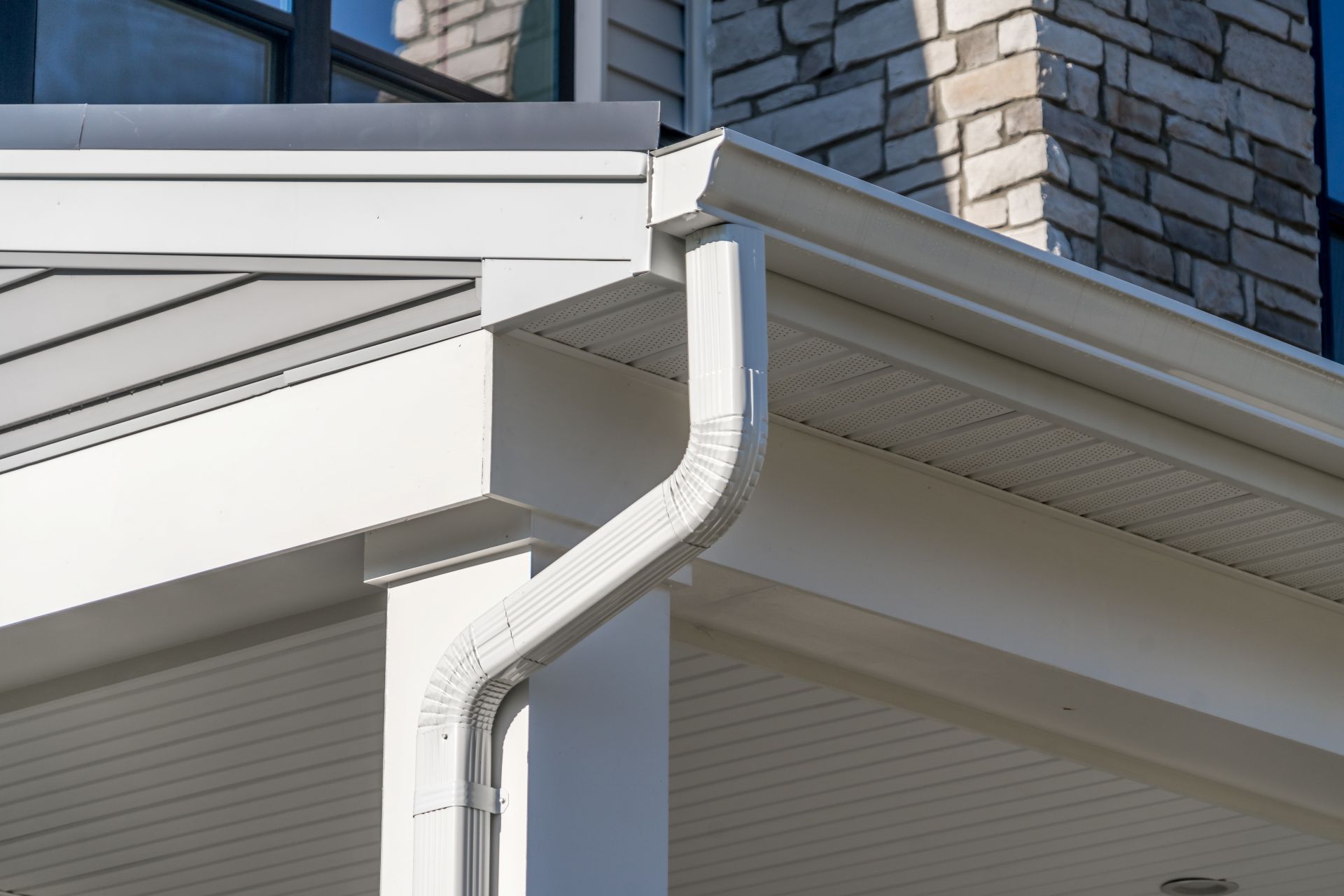 White gutter and downspout on a house corner beneath a stone chimney and soffit