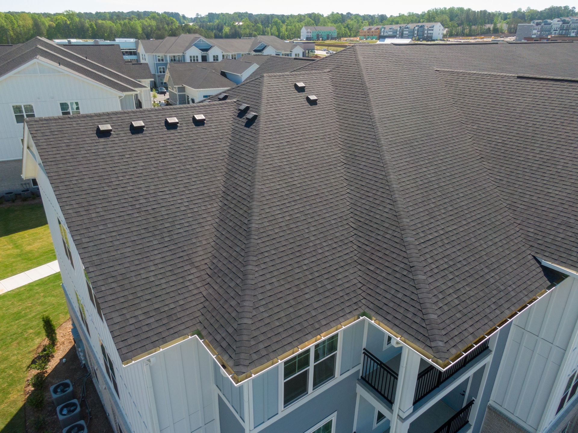 Aerial view of a gray shingle apartment roof with multiple peaks and vents in a residential neighborhood