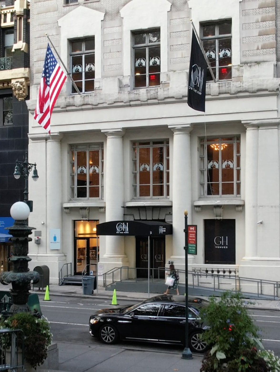 White stone building with American and black flags, black awning, and parked black car on a city street.