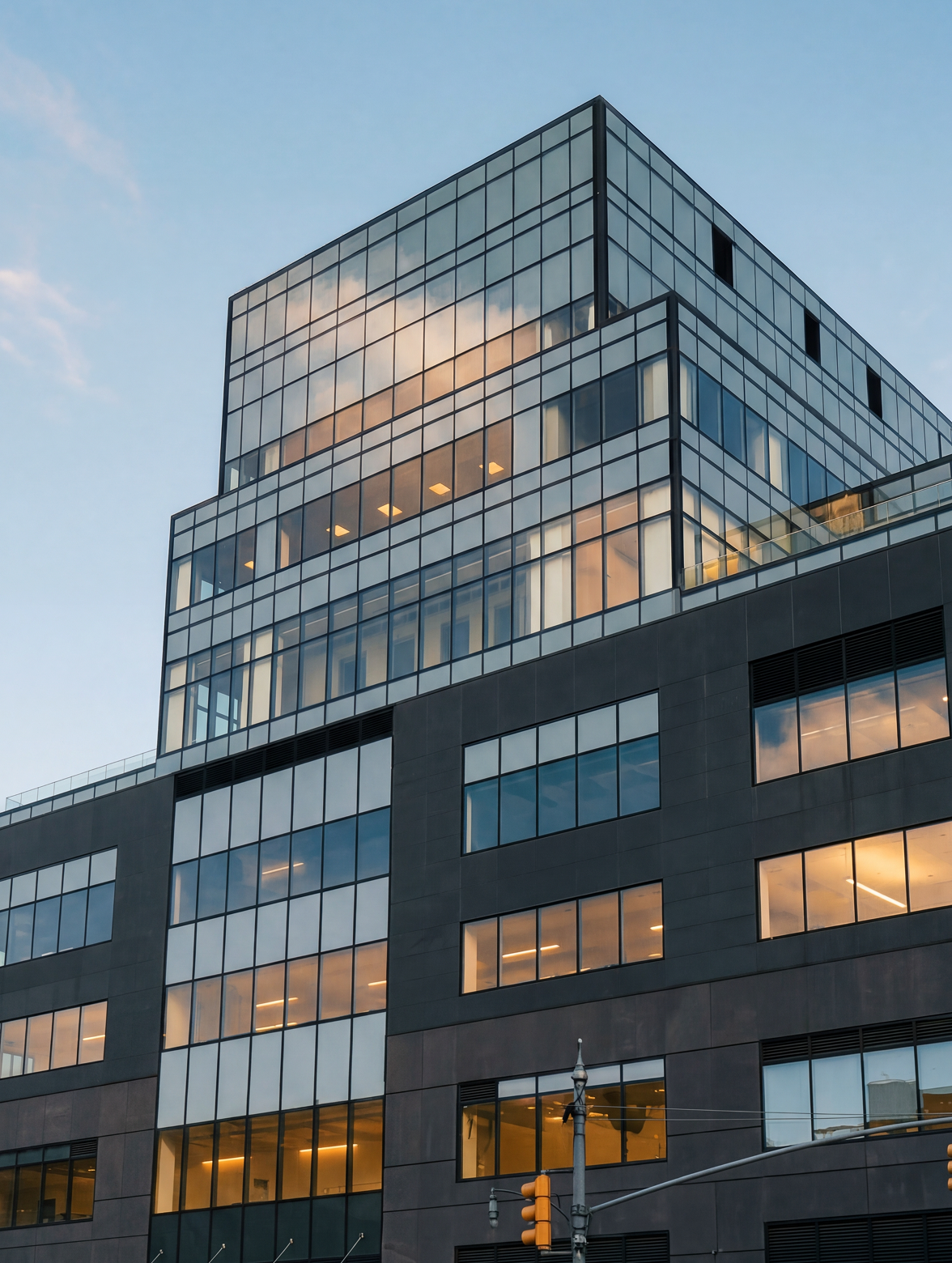 Modern glass office building at dusk with warm-lit windows and a cloudy sky