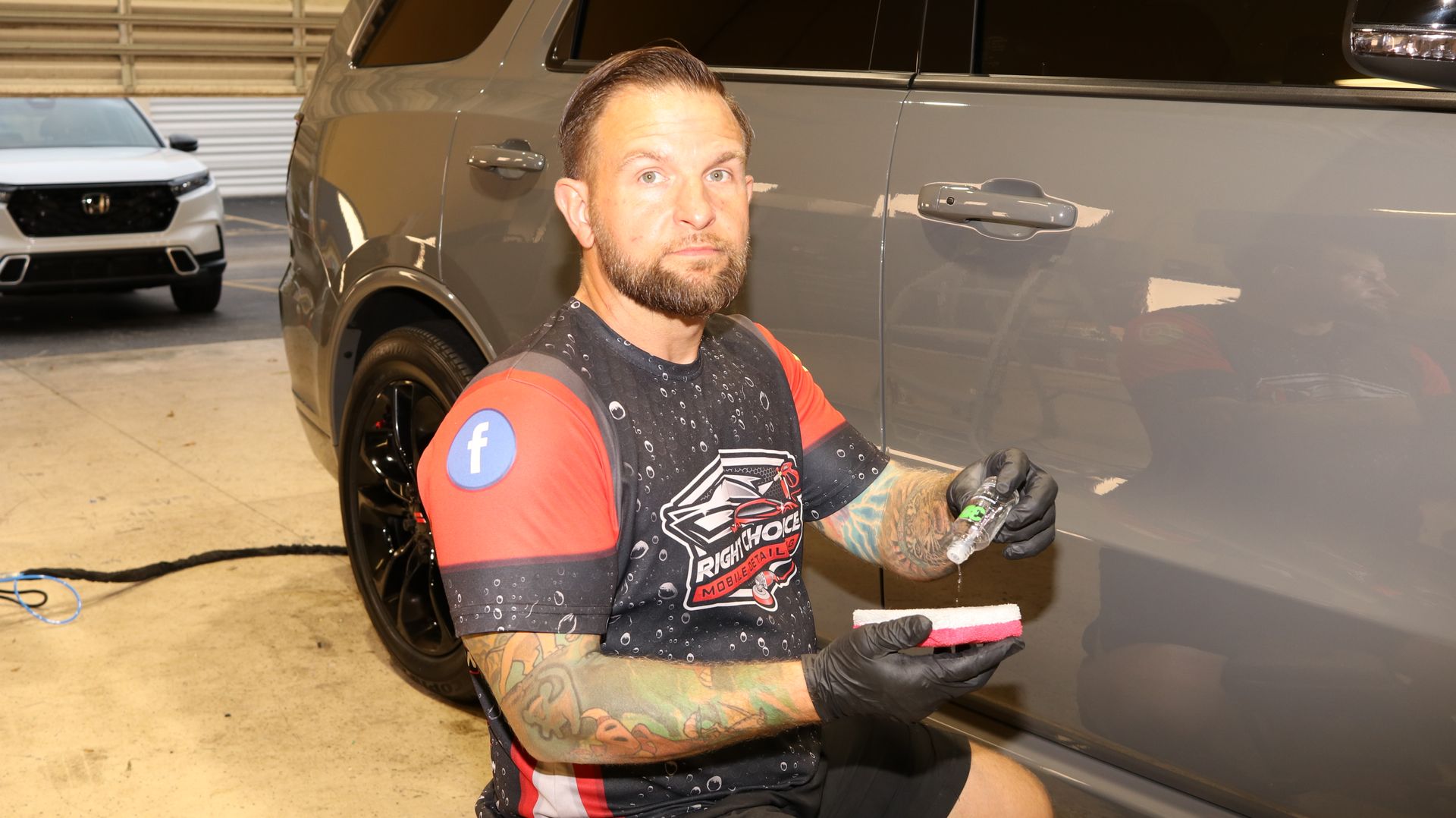 Man applying detailing product to gray SUV's side. He's kneeling and wearing black gloves. Car is in a garage.