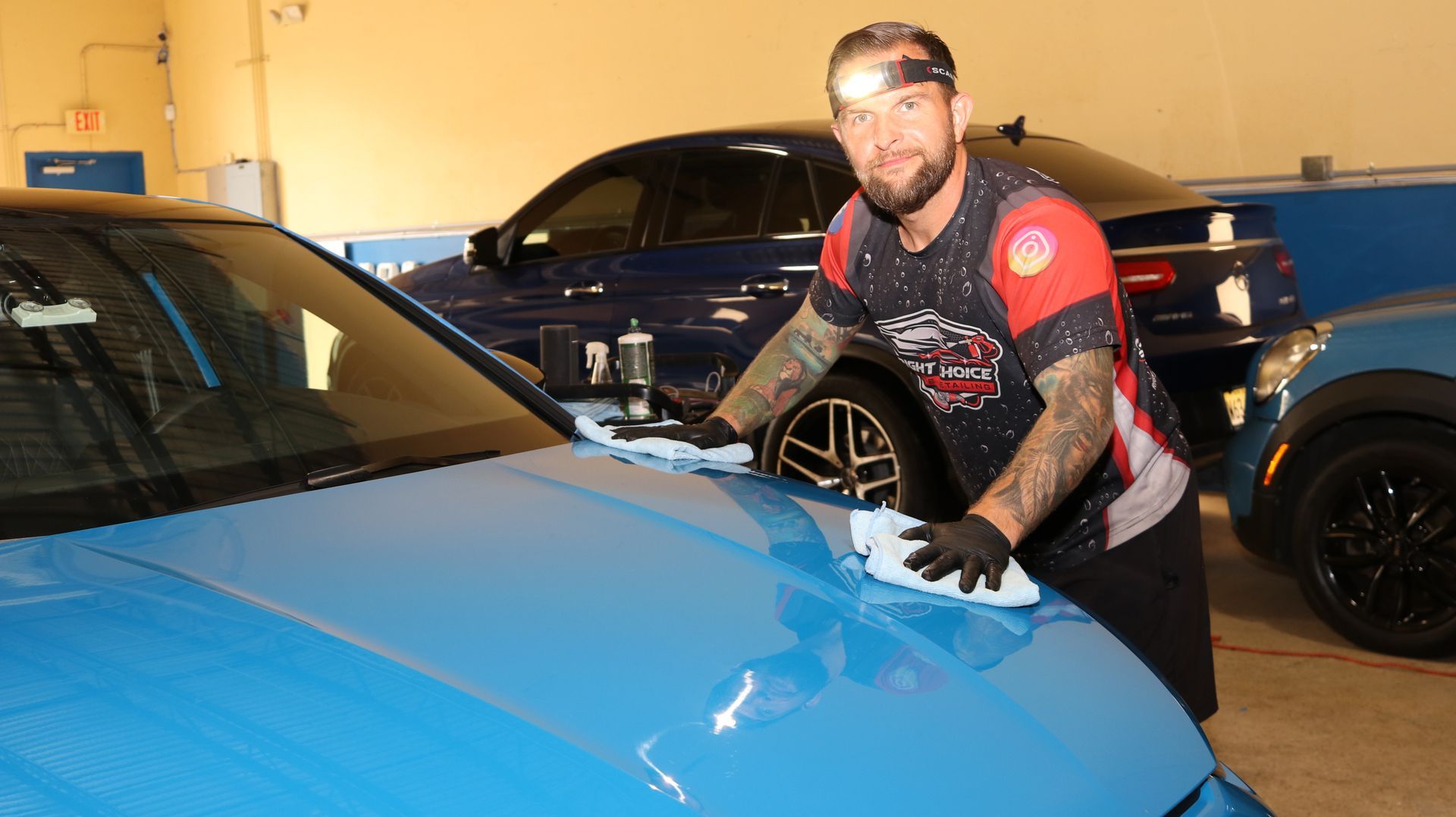 Man cleaning the hood of a blue car. He wears gloves, a headlamp, and has tattoos. Other cars in background.