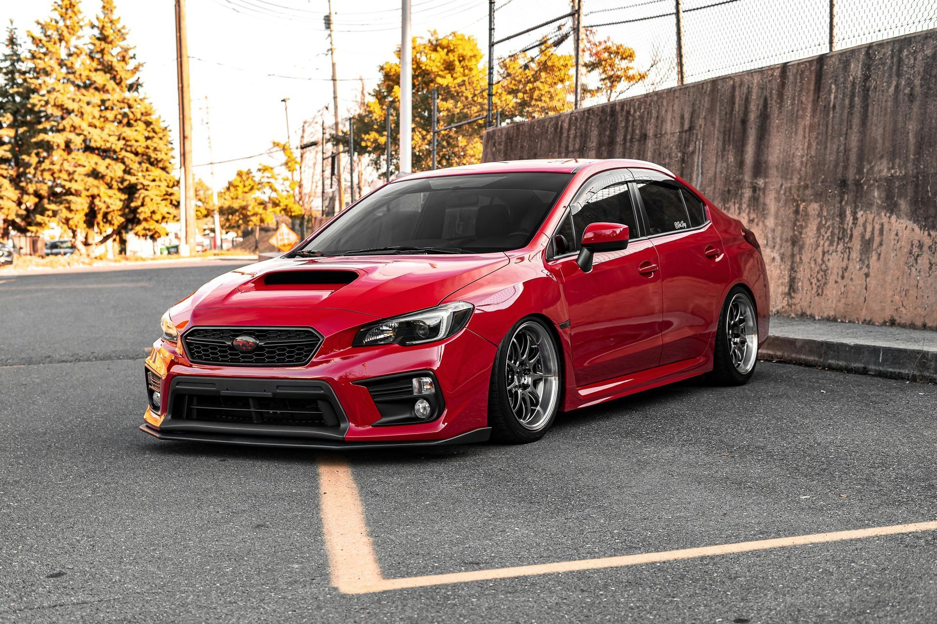Red sports sedan parked on a street beside a concrete wall and autumn trees