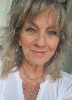 Close-up portrait of a smiling woman with wavy gray hair wearing a white blouse and layered necklaces.