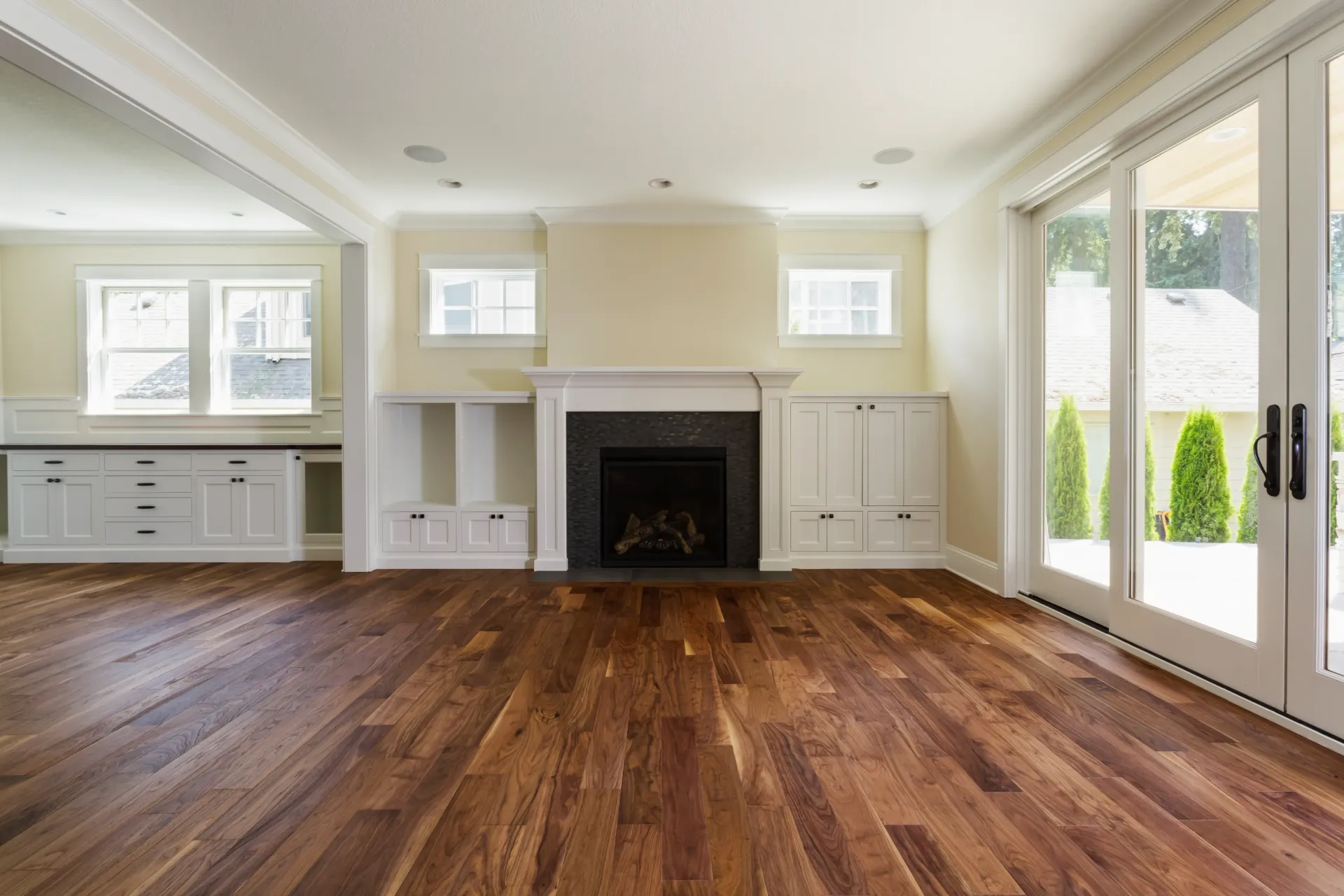 Bright empty living room with hardwood floors, white fireplace, and large windows/doors opening to greenery