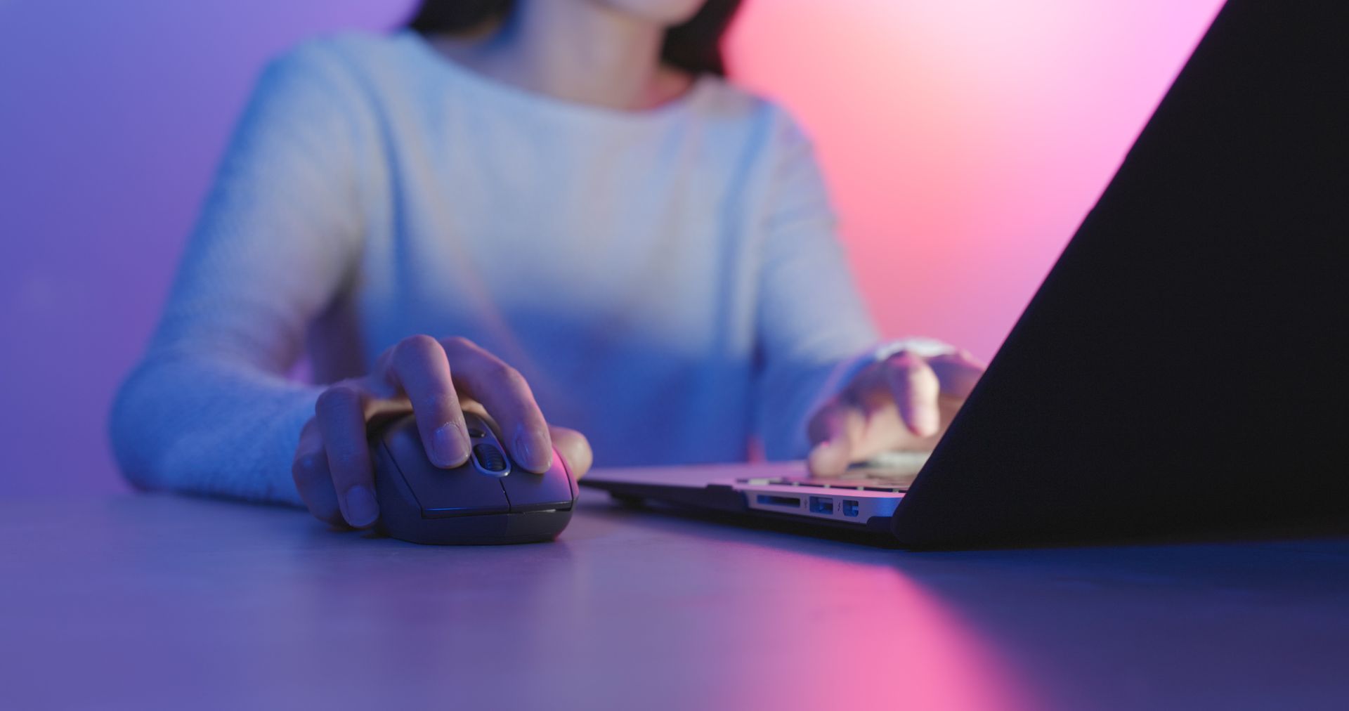Woman using a laptop, holding a mouse. The scene is lit with purple and pink light.