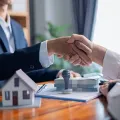 Two people shake hands over a table with a house model, a stack of money, and a document stamp.