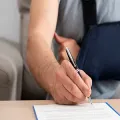 A person with their arm in a blue sling sits at a table signing a document with a black pen.