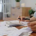 A person sits at a wooden desk with a calculator and financial papers, using a pen to manage budget documents.