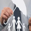 A business person holds a paper cutout of a family under a paper umbrella, symbolizing insurance and protection.