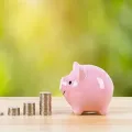 A pink piggy bank sits on a wooden surface next to four stacks of coins of increasing height against a green background.