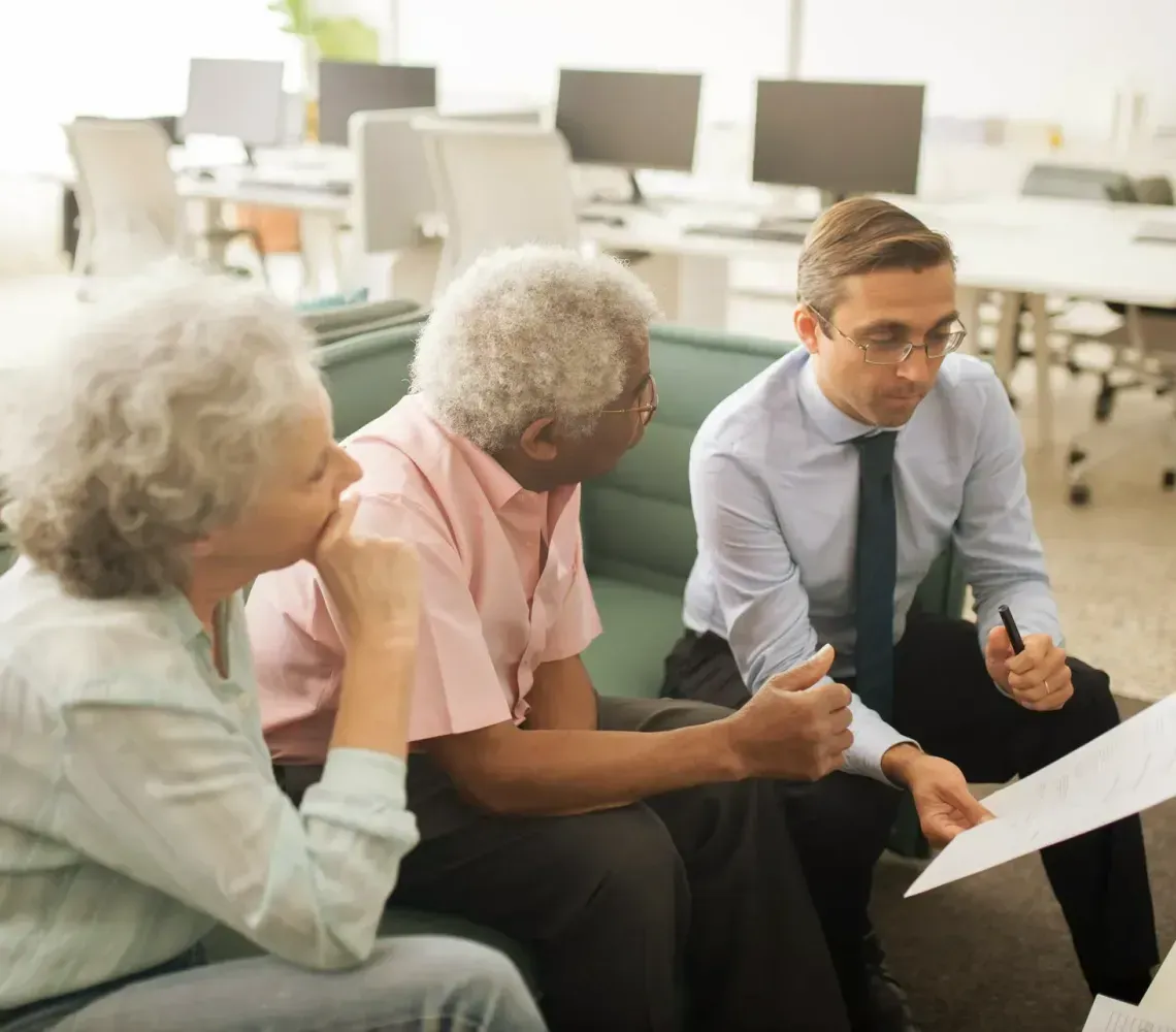 A professional holds a document while discussing information with two people seated on a couch in an office setting.