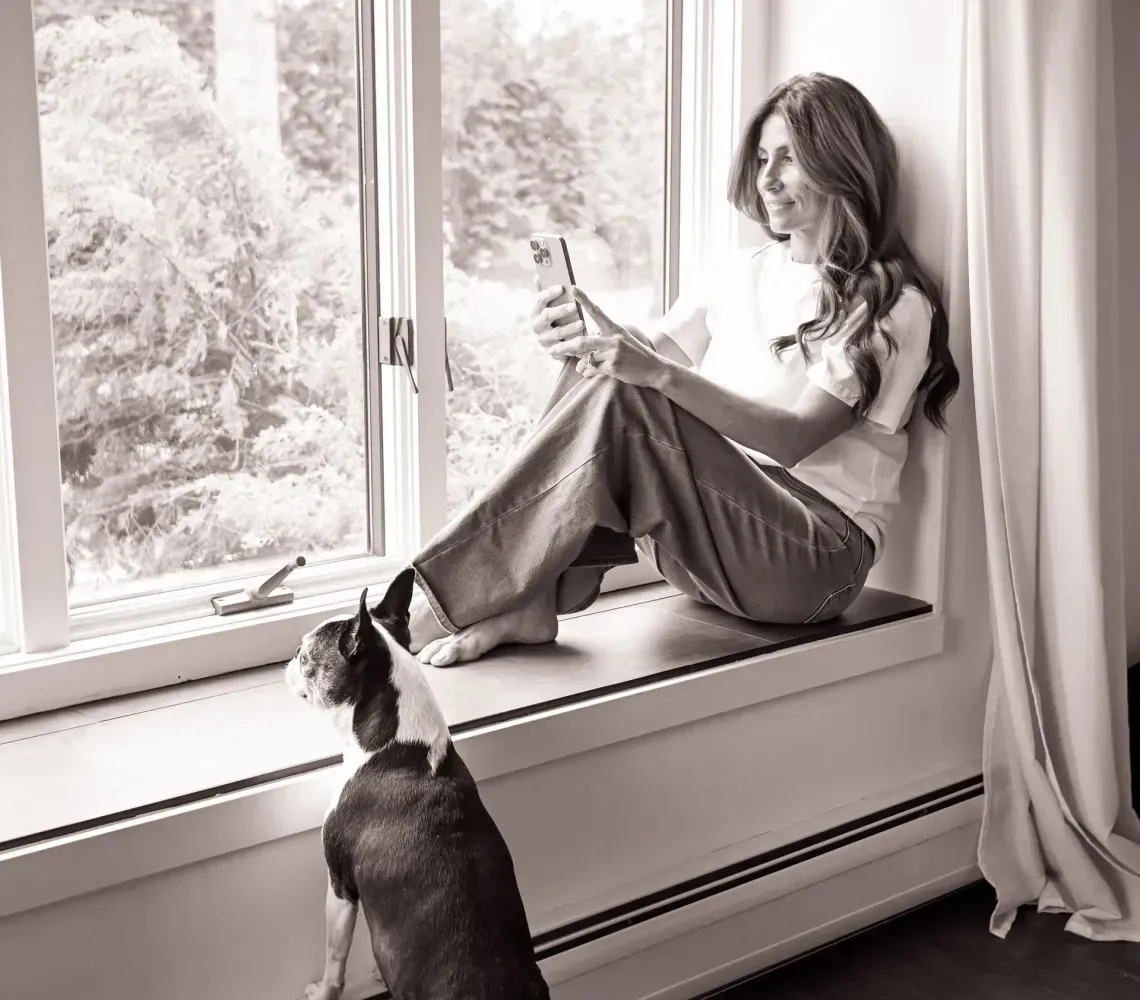 A person sits on a window ledge looking at a smartphone, while a dog stands on the floor looking out the window.