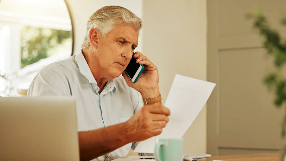 A person in a light shirt talks on a smartphone while holding a document at a desk with a laptop and coffee mug.
