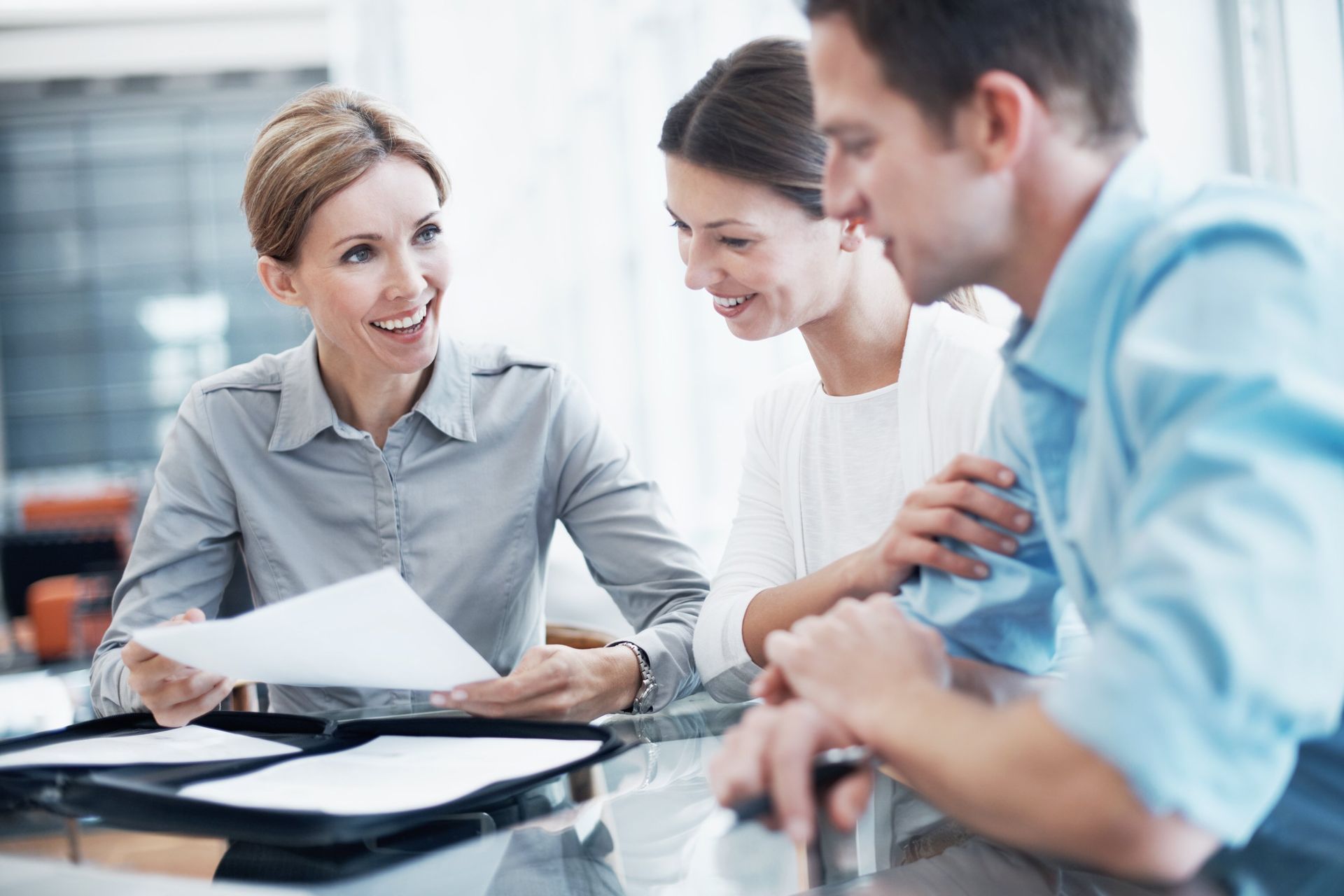 A professional meeting where a smiling person reviews documents with a couple at a desk in a bright office.