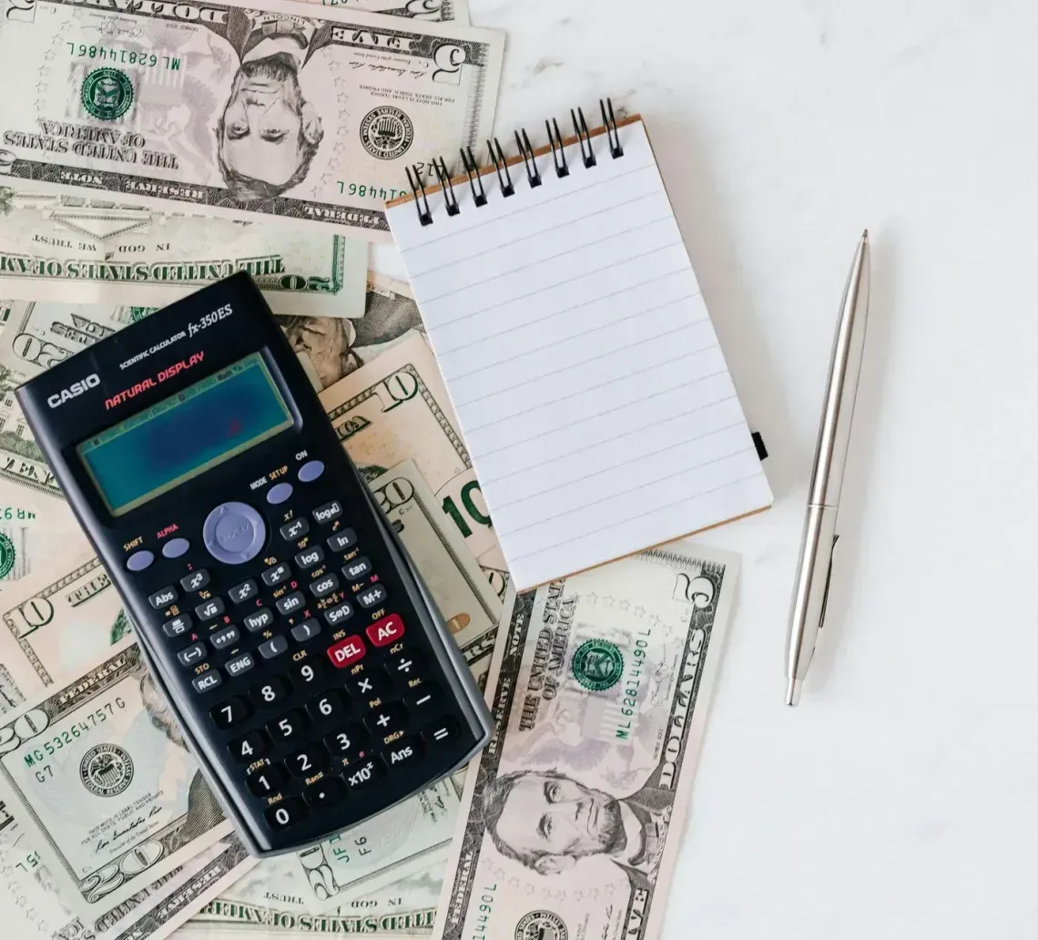 Calculator, notepad, and pen on a pile of scattered US dollar bills, on a white surface.