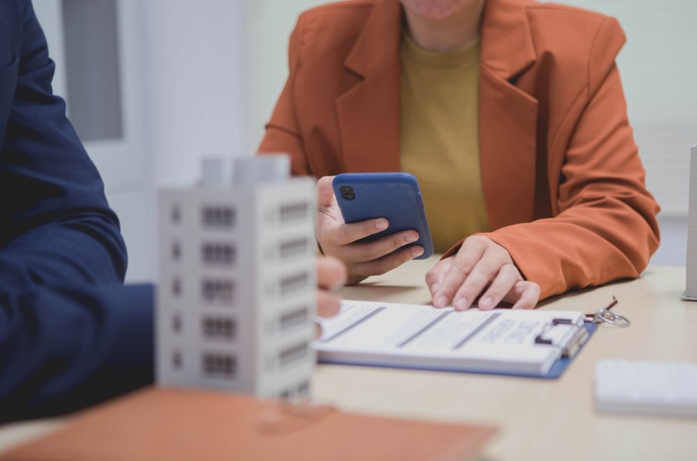 Person in orange blazer using phone at a table with documents and building model.