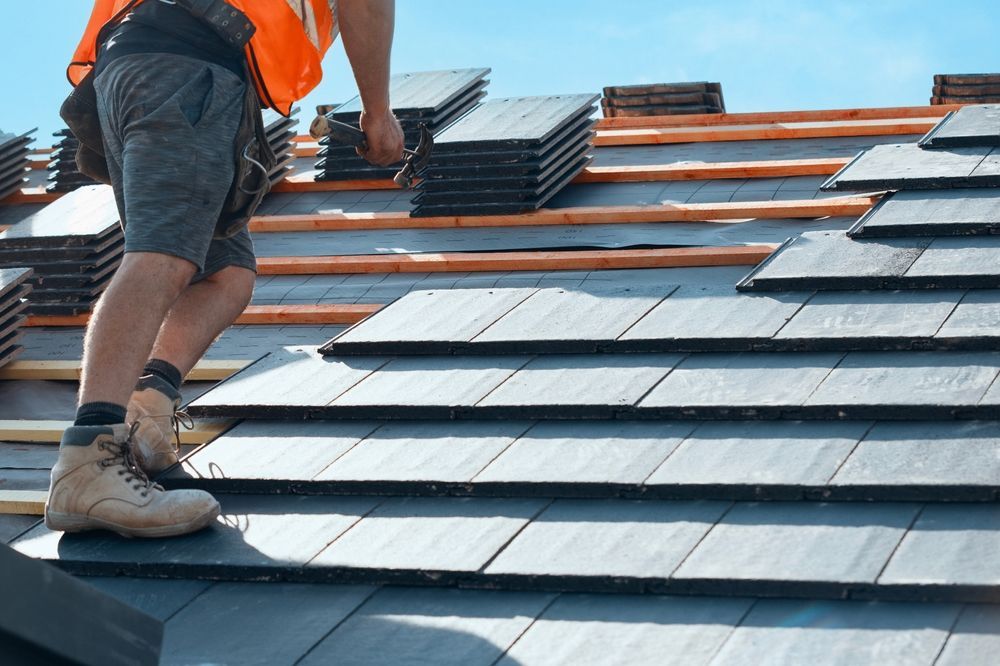 Roofer installing dark gray tiles on a roof, sunny day.